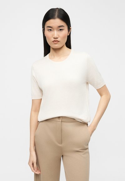 Γυναίκα με μακριά σκούρα μαλλιά φοράει λευκό κοντομάνικο τοπ και μπεζ παντελόνια, στέκεται με το ένα χέρι στην τσέπη μπροστά σε απλό φόντο.