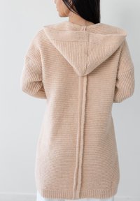 Beżowy sweter z dzianiny z kapturem, z teksturowanym wzorem, otwarty z przodu i z szwem z tyłu. Długie rękawy i ściągacze na mankietach.