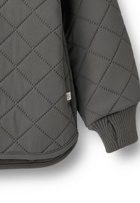 Tikkitakki harmaana, jossa on timanttikuvio, resorit hihansuissa ja pieni brändätty merkki alaosassa. Pehmeä, teksturoitu materiaali.