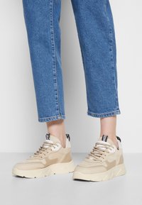Beżowe sneakersy z połączeniem gładkich i zamszowych tekstur, masywną podeszwą, sznurowanym designem oraz czarną metką na pięcie.