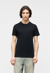 Νέος άνδρας με μεσαίου μήκους σκούρα μαλλιά, φορώντας απλή μαύρη κοντομάνικη μπλούζα και ανοιχτόχρωμο μπλε τζιν, στέκεται μπροστά σε λευκό φόντο.