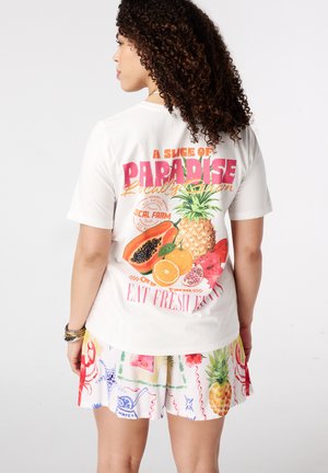 Vrouw met krullend haar die een wit shirt draagt met een kleurrijke tropische fruitprint en tekst, gecombineerd met witte korte broek met verschillende fruit- en zeemotieven.