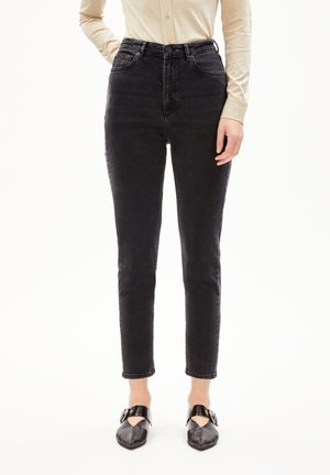 Femme portant un jean slim noir taille haute, un haut beige à manches longues et des chaussures pointues en cuir noir avec des brides à boucle.