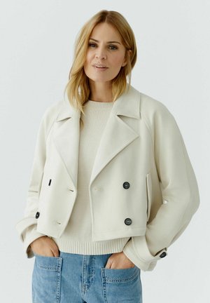 Blonde Frau, die eine cremefarbene, kurz geschnittene, doppelt geknöpfte Jacke über einem weißen Pullover und blauen Jeans trägt, steht mit den Händen in den Taschen vor einem schlichten Hintergrund.