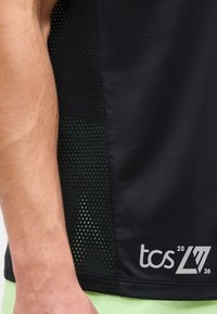 Χέρι και πλευρό ενός ατόμου που φοράει μαύρο αθλητικό πουκάμισο από δίχτυ με λογότυπο "tcs 20 26" κοντά στο τελείωμα και ανοιχτό πράσινο παντελόνι.