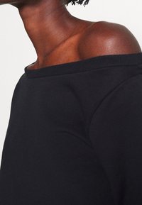 Top negro sin mangas con escote tipo off-the-shoulder y cuello acanalado, confeccionado en una tela suave y texturizada. Presenta un corte holgado que destaca la zona de la clavícula.