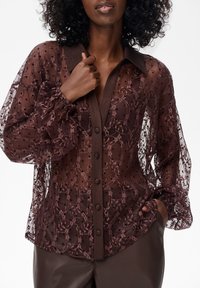 Przezroczysta, ciemnobrązowa bluzka z wzorzystą koronką i kropkami. Posiada szeroki kołnierz, zapięcie na guziki z przodu oraz marszczone mankiety.