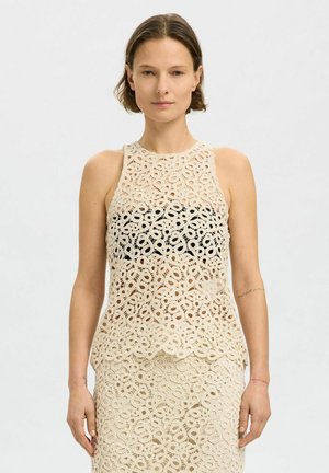 Hækelagt tanktop i beige med en blonde mønster og bølget kant, med et sort underlag for ekstra kontrast.