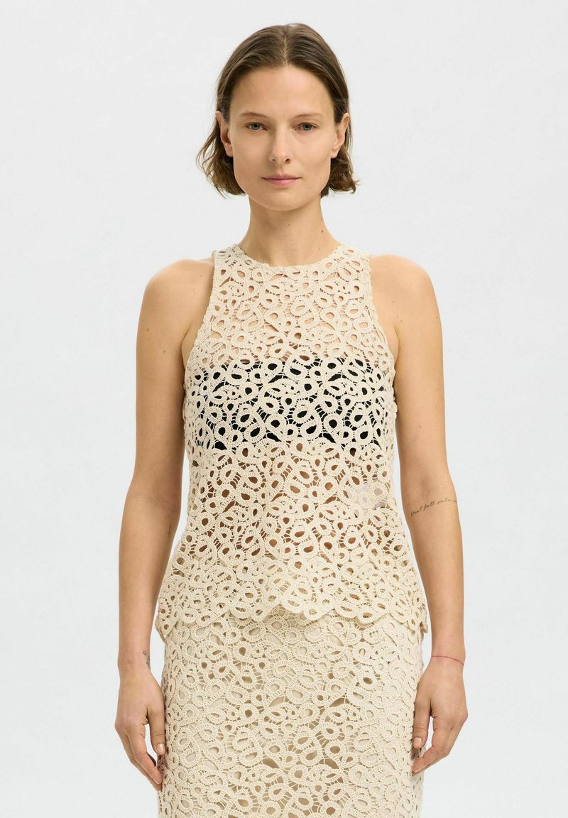 Hækelagt tanktop i beige med en blonde mønster og bølget kant, med et sort underlag for ekstra kontrast.