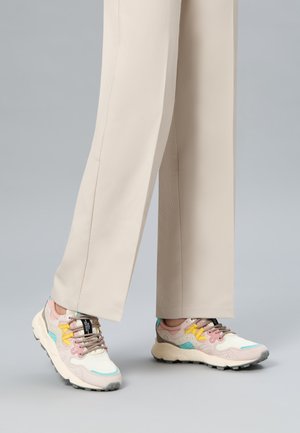 Persona che indossa pantaloni beige a gamba larga e sneakers multicolore pastello con suole bianche su pavimento grigio.