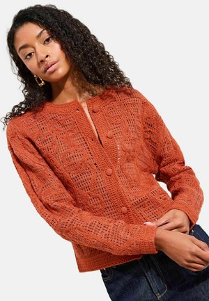 Frau mit lockigem Haar, die eine orangefarbene Strickjacke mit Struktur und Knöpfen trägt, kombiniert mit dunkelblauer Jeans, vor weißem Hintergrund.