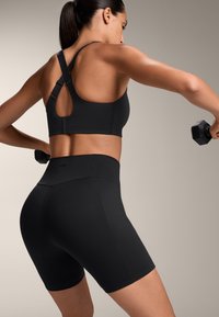 Női fekete sportmelltartó racerback dizájnnal, magas derékú fekete rövidnadrággal párosítva, mindkettő sima, rugalmas anyagból készült.