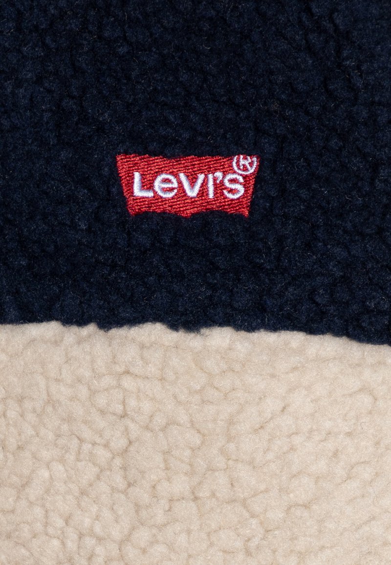 Chaqueta de tejido polar de Levi's, con una sección superior en azul marino y una inferior en color crema. Logo bordado en rojo prominentemente exhibido.