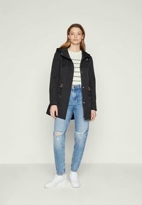 Mulher em pé, vestindo uma jaqueta preta com capuz sobre um suéter branco estampado, jeans claros rasgados e ténis brancos, contra um fundo simples.