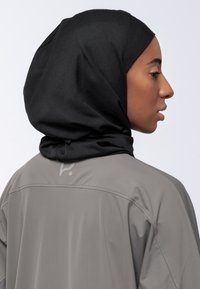 Donna di profilo che indossa un hijab nero e una giacca grigia con un logo sottile sulla schiena, su uno sfondo bianco uniforme.