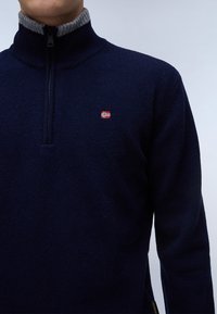 Granatowy sweter dzianinowy z wysokim kołnierzem, szarymi ściągaczami, ćwierć zamkiem błyskawicznym i małą haftowaną norweską flagą na piersi.