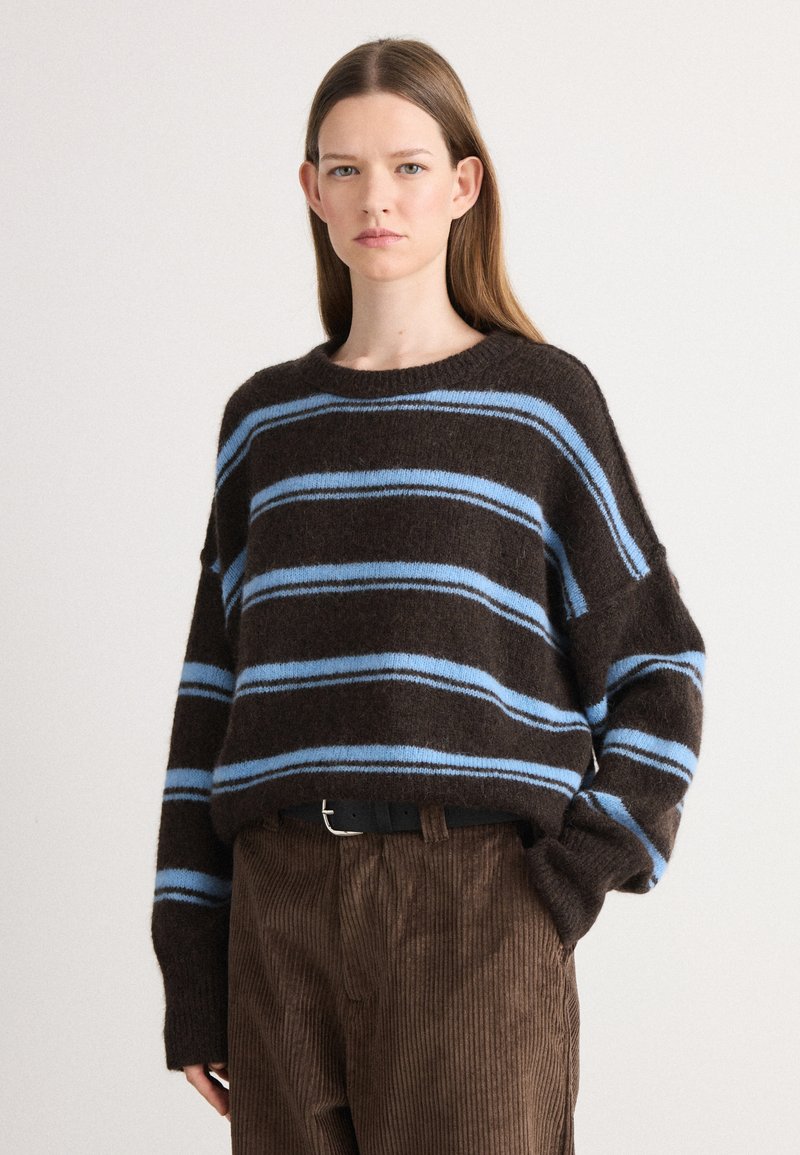 Braun gestreifter Pullover mit blauen Horizontalstreifen, aus einem strukturierten Strickmaterial. Getragen mit braunen Cordhosen und einem schwarzen Gürtel.