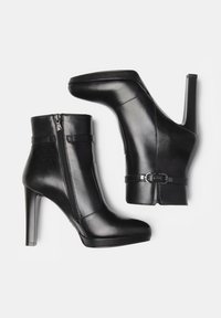 Black leather ankle boots with a pointed toe, high stiletto heel, side zipper, and buckle accents. Smooth texture with a sleek design.