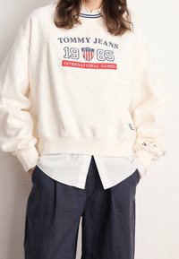 Kremowy, krótki sweatshirt z niebieskim i czerwonym napisem, z okrągłym dekoltem i ściągaczami na rękawach, nałożony na białą koszulę z guzikami.
