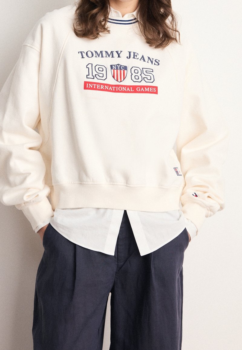 Kremowy, krótki sweatshirt z niebieskim i czerwonym napisem, z okrągłym dekoltem i ściągaczami na rękawach, nałożony na białą koszulę z guzikami.
