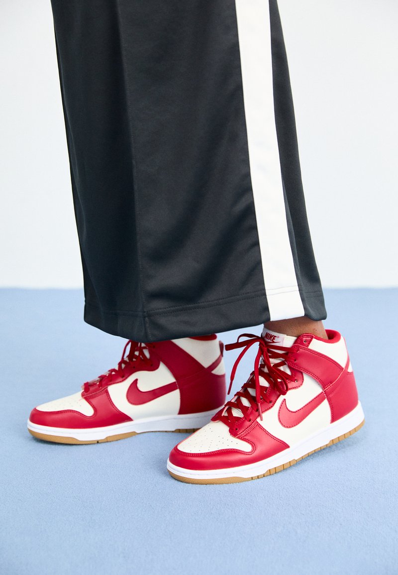 Personne portant un pantalon noir avec une rayure blanche et des baskets montantes Nike rouge et blanche se tenant sur un sol bleu.