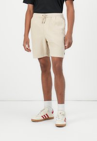 Beige joggingshorts met een gestructureerde stof, zijzakken en een relaxte pasvorm. Gepaard met witte sneakers met rode strepen.