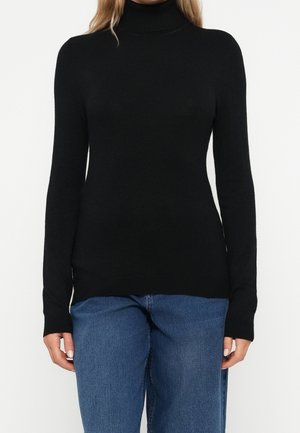 Femme portant un pull à col roulé noir moulant à manches longues, associé à un jean bleu, debout devant un fond blanc uni.