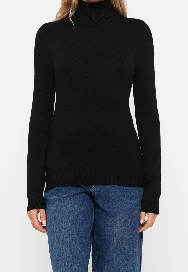 Femme portant un pull à col roulé noir moulant à manches longues, associé à un jean bleu, debout devant un fond blanc uni.