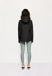 Giacca impermeabile nera con cappuccio, abbinata a leggings verde marmorizzati e sneakers multicolori, vista da dietro.