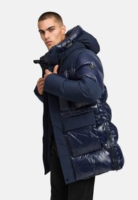 Laivansetin puffatakki, jossa on kiiltävä pinta, suuri huppu, useita vetoketjullisia taskuja ja joustavat hihansuut lämpöä ja mukavuutta varten.