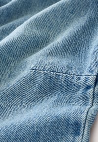 Jasnoniebieska tkanina dżinsowa o subtelnej fakturze, z małą naszytą kieszonką i widocznymi detalami szwów. Miękka i lekko pomarszczona.