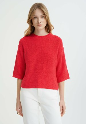 Red knitted short-sleeve sweater with a round neckline, textured finish, and a relaxed fit, paired with white pants.