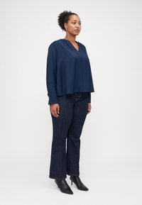 Mörkblå, lättviktig blus med lång ärm och V-ringning, med en lätt drapering, tillsammans med mörka denimjeans med utsvängda ben och svarta spetsiga stövlar.