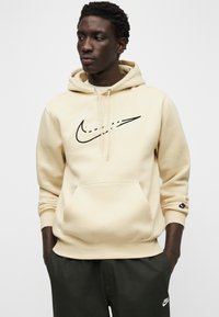 Bež Nike jopa s črnim logotipom na prsih. Ima žep kenguru, kapuco z vrvico in rebraste rokave. Nosi se s temno zelenimi hlačami.