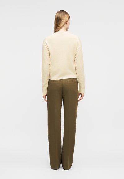 Γυναίκα που στέκεται πλάτη, φοράει κρεμ πουλόβερ με μακριά μανίκια και ελιά φαρδιά παντελόνια σε απλό φόντο.