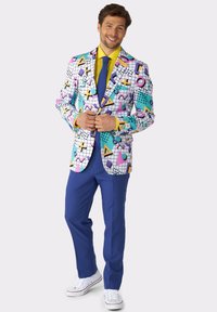 Giacca da uomo colorata con motivi geometrici in rosa, blu e giallo, abbinata a pantaloni blu e scarpe bianche. Camicia gialla a maniche lunghe.