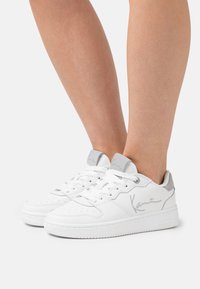 Hvite lær sneakers med sølvdetaljer, med perforert tå, snøring og polstret krage. Signaturlogo synlig på siden.
