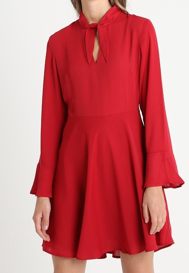 Robe rouge à manches longues avec jupe évasée et encolure keyhole nouée, portée par une personne les mains détendues le long du corps.