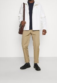 Mężczyzna w białej kurtce, granatowej koszuli, beżowych spodniach, czarnych sneakersach i trzymający brązową skórzaną torbę na ramieniu, stojący na jednolitym tle.