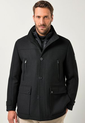 Uomo che indossa un cappotto invernale nero con bottoni, dotato di tasche con zip e pattina, pantaloni beige e uno strato interno trapuntato nero su uno sfondo semplice.