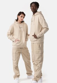 Beige hættetrøjer med forlommer og snoretræk, parret med beige cargo bukser med flere lommer. Begge har hvide sneakers på.