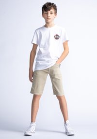 Witte katoenen t-shirt met een rond logo-embleem, lichtbeige shorts en witte hoge sneakers. Model poseert met één hand in de zak.