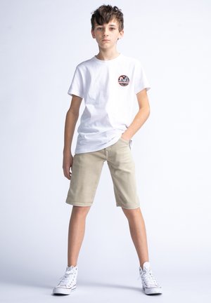 T-shirt en coton blanc avec un écusson rond, shorts beige clair et baskets montantes blanches. Le mannequin pose avec une main dans la poche.