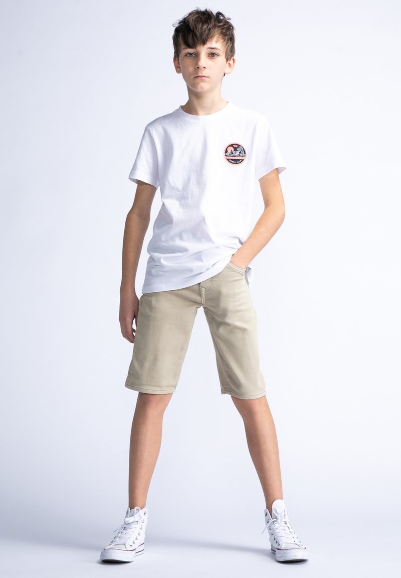 Witte katoenen t-shirt met een rond logo-embleem, lichtbeige shorts en witte hoge sneakers. Model poseert met één hand in de zak.