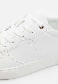Zapatilla de cuero blanco con cordones planos blancos, ojales con acentos dorados y detalles perforados en el costado. Suela de goma plana.