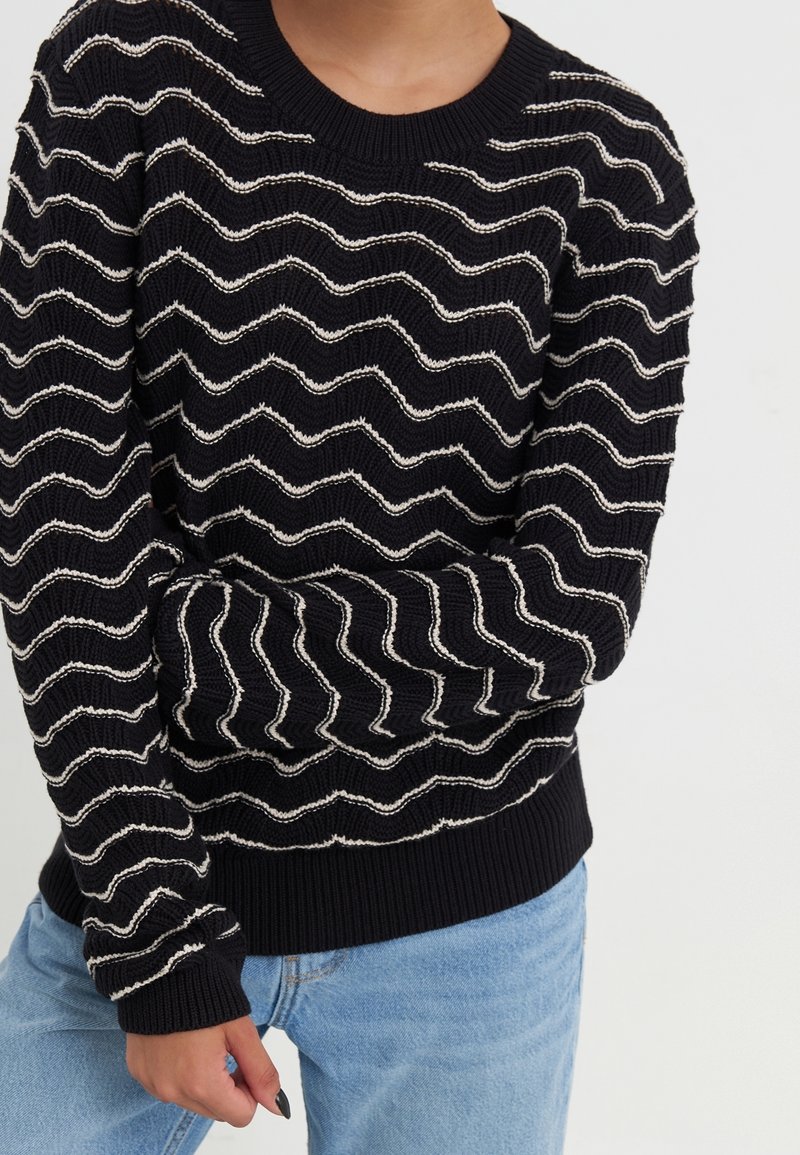 Personne portant un pull en maille noir à rayures blanches ondulées et un jean bleu clair, les bras croisés à la taille.