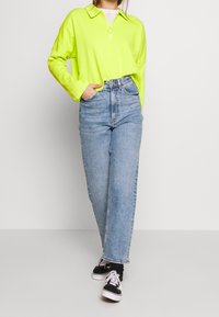 Polo shirt a maniche lunghe verde neon con colletto zip, abbinato a jeans azzurri a vita alta e sneaker nere con suola bianca.