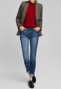 Brunternetternetjakke over en rød rullekrave, parret med blå denimjeans rullet op ved stængerne og sorte slip-on sko.