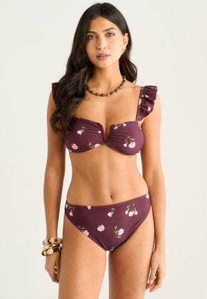 Woman with long dark hair wears a purple floral bikini with ruffled shoulder straps and matching bottoms against a plain light background.