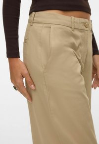 Cilvēks, valkājot bēšas augsta vidukļa bikses ar sānu kabatām, kopā ar tumšu garu piedurkņu kreklu, rādot roku ar gredzenu, kas atpūšas pie kabatas.